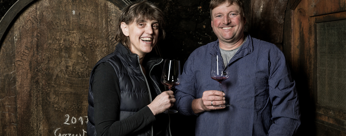 Zwei Personen mit Weingläsern lächelnd vor einem großen Holzfass.