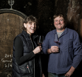 Zwei Personen mit Weingläsern lächelnd vor einem großen Holzfass.
