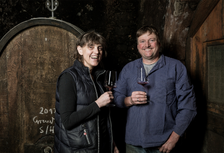 Zwei Personen mit Weingläsern lächelnd vor einem großen Holzfass.