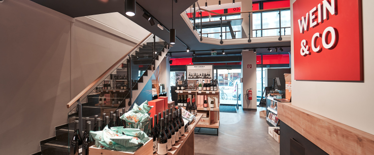 Moderner Weinladen mit offenem Grundriss, Holztischen und Treppe im Hintergrund.