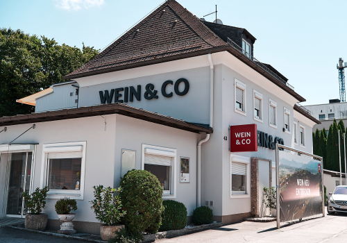 Graues Gebäude mit braunem Dach und weinrotem Schild, umgeben von Pflanzen.