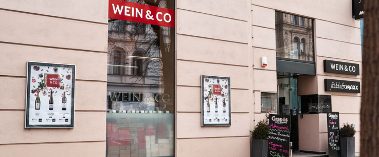 Eingang eines modernen Geschäfts mit großen Fenstern und Werbeplakaten an der Fassade.