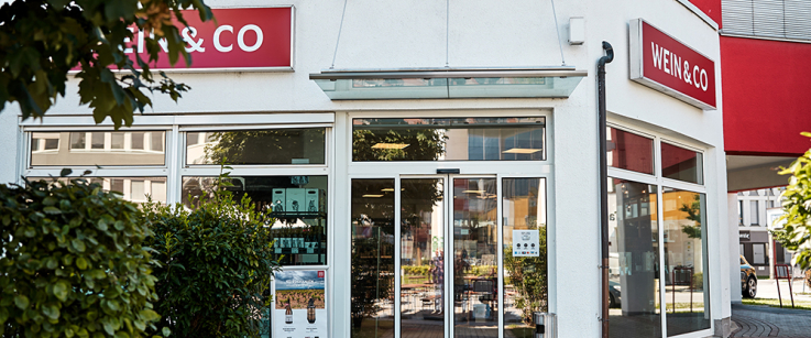 Moderner Laden mit Glasfront und roten Fassadenschildern, umgeben von Bäumen und Sträuchern.