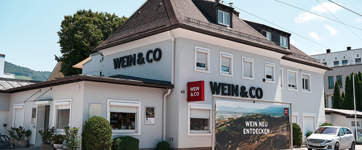 Grau-weißes Gebäude mit Werbetafel und Fensterläden, davor parkt ein silbernes Auto.