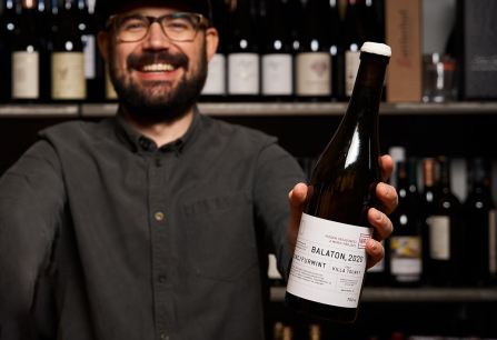 Person präsentiert eine Weinflasche vor einem Regal mit weiteren Weinflaschen.