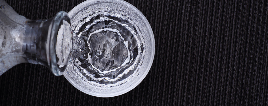 Karaffe gießt Wasser in Glas auf schwarzem, texturiertem Hintergrund.