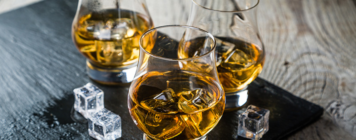 Drei Whiskygläser mit Eiswürfeln auf dunklem Tablett, rustikaler Holztisch.