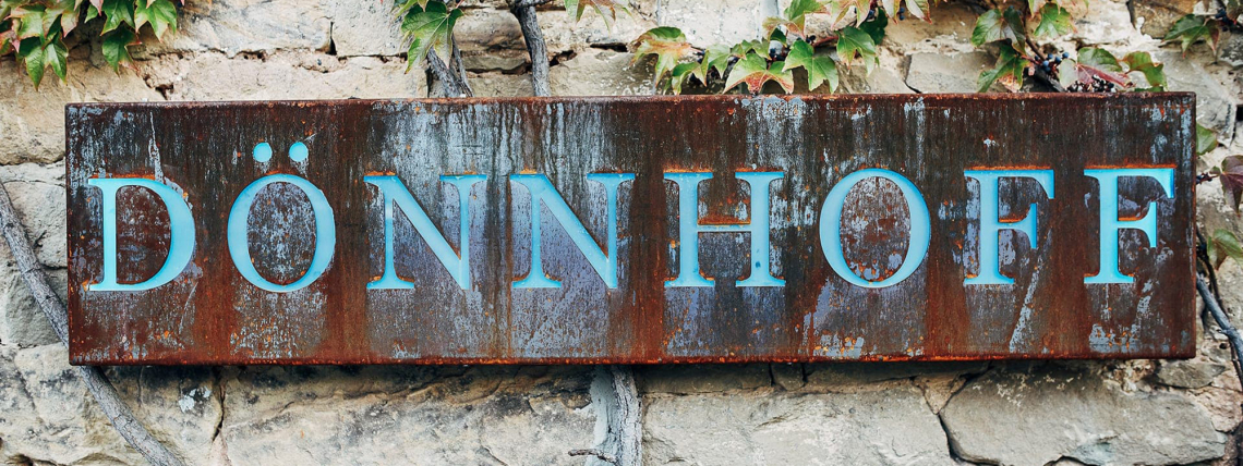 Rostiges Metallschild an einer Steinwand mit blauen Buchstaben, umgeben von grünen Blättern.