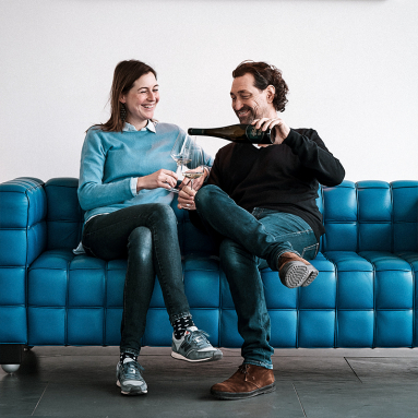 Zwei Personen auf blauer Couch, lachen und schenken Wein ein.