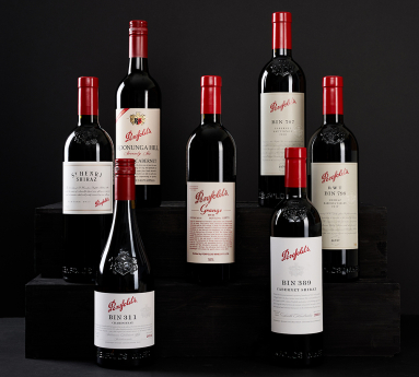 Sieben Weinflaschen mit roten Kapseln und eleganten Etiketten auf schwarzen Holzregalen.