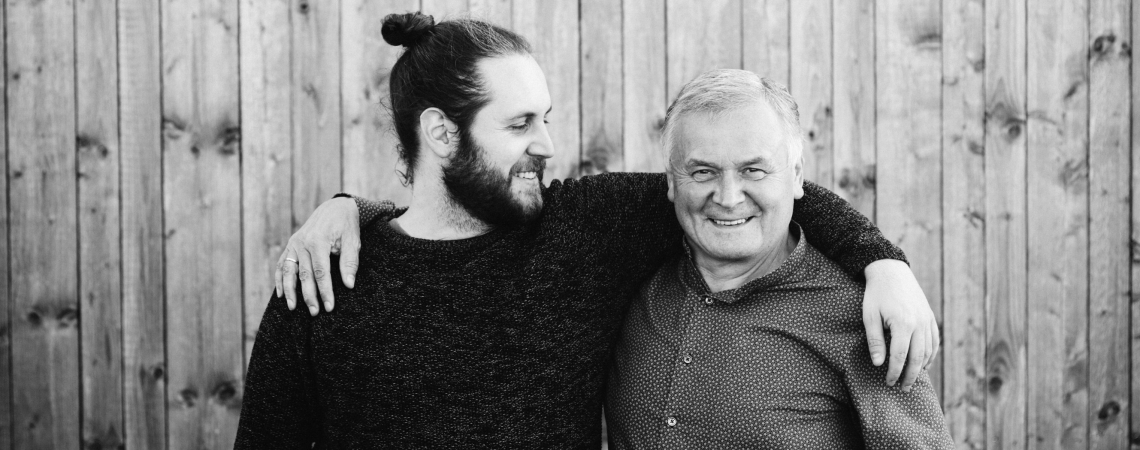 Zwei Personen lächeln vor Holzwand, freundschaftlich Arm um Schulter gelegt.