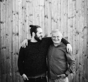 Zwei Personen lächeln vor Holzwand, freundschaftlich Arm um Schulter gelegt.