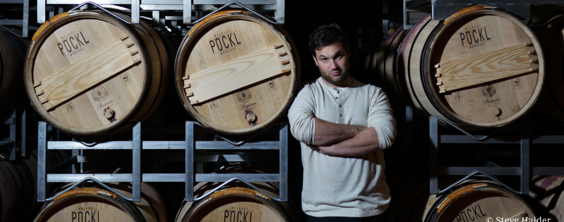 Mann in hellem Pulli lehnt an Holzfässern in dunklem Weinkeller.