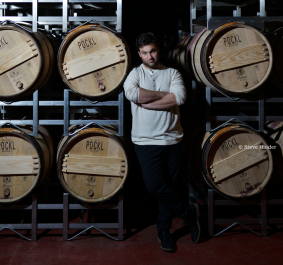 Mann in hellem Pulli lehnt an Holzfässern in dunklem Weinkeller.