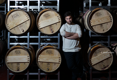 Mann in hellem Pulli lehnt an Holzfässern in dunklem Weinkeller.