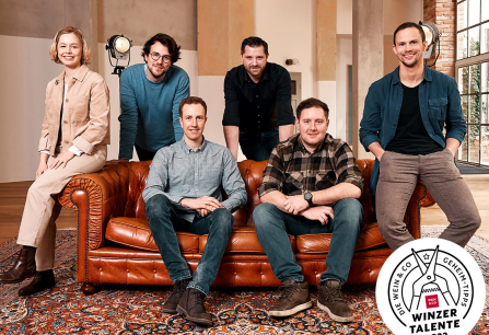 Sechs Personen sitzen und stehen um ein braunes Ledersofa in einem Loft-Raum.