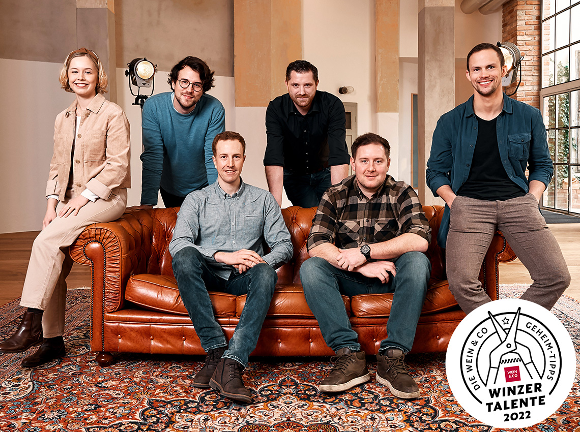 Sechs Personen sitzen und stehen um ein braunes Ledersofa in einem Loft-Raum.