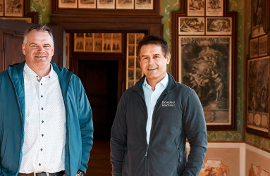 Zwei Männer in einem mit Gemälden dekorierten, historischen Raum.