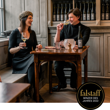 Zwei Personen trinken Wein an einem Holztisch in stilvoller Umgebung mit Weinflaschen.