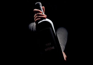 Dunkle Weinflasche in Händen vor schwarzem Hintergrund.