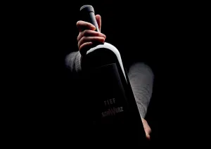 Dunkle Weinflasche in Händen vor schwarzem Hintergrund.