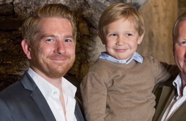 Drei Generationen lächeln vor rustikaler Steinwand in eleganter Kleidung.