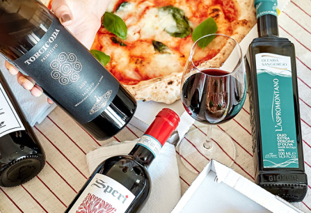 Flaschen Wein und Olivenöl neben Pizza auf gestreiftem Tischtuch.