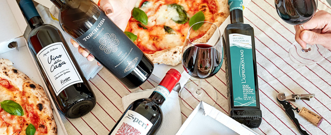 Flaschen Wein und Olivenöl neben Pizza auf gestreiftem Tischtuch.