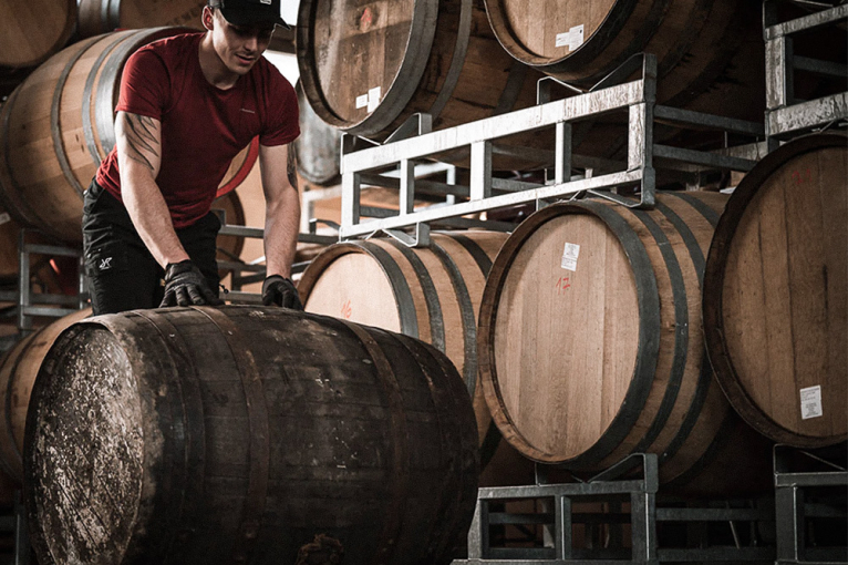 Mann bewegt großes Holzfass in einer Lagerhalle voller Fässer.