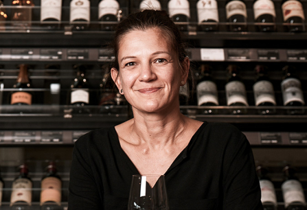 Frau in schwarzem Oberteil vor einem Weinregal, hält ein Weinglas.
