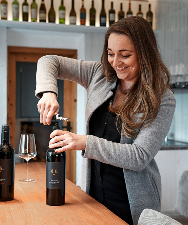 Frau öffnet Weinflasche in modernem Innenraum, lächelnd an Holztresen.